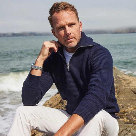 A smiling man in sunglasses wears the COLLARS AND CO - PACIFIC 1/4 ZIP COTTON SWEATER IN NAVY outdoors by the water, with a bridge and rocky shoreline in the background.