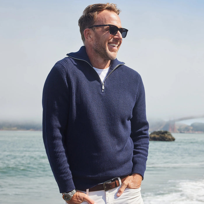 A man wearing the COLLARS AND CO - PACIFIC 1/4 ZIP COTTON SWEATER IN NAVY and white pants sits on rocks by the ocean, gazing thoughtfully at the misty coastline with waves in the background.