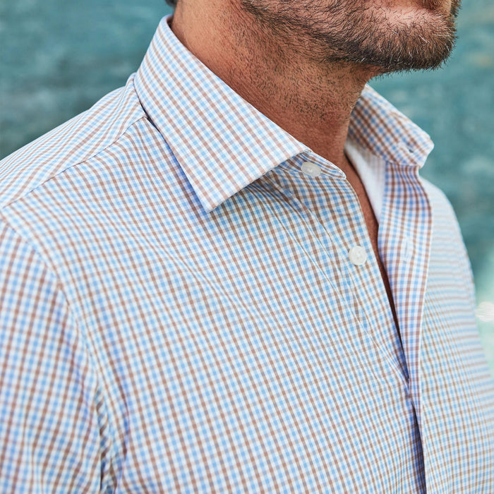 A man with short brown hair and a beard, wearing the COLLARS AND CO Semi Spread Collar Apex Performance Dress Shirt in Tan and Blue Tattersall, sits on a bench with his hands clasped, looking to the side by a blue textured wall.