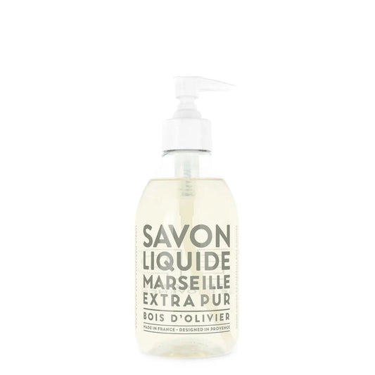 A clear pump bottle of COMPAGNIE DE PROVENCE Liquid Marseille Soap Olive Wood 10oz by CIE LUXE BRANDS, with a light-colored soap and a white pump dispenser. White label text reads "Savon Liquide Marseille Extra Pur Bois d’Olivier.