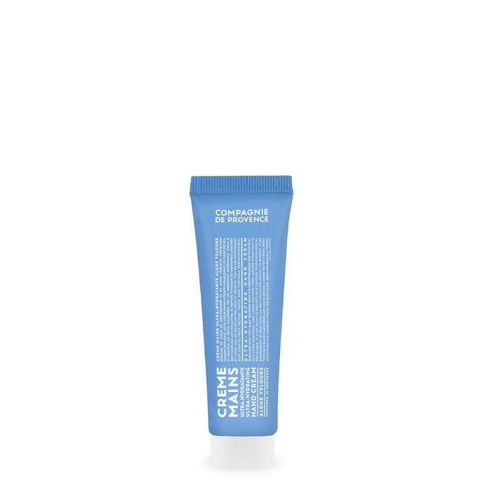 A blue tube of CIE LUXE BRANDS COMPAGNIE DE PROVENCE - TRAVEL HAND CREAM VELVET SEAWEED 10Z stands next to a beaker of water and green seaweed, set against a dark blue background to showcase its botanical oils for lasting hydration.