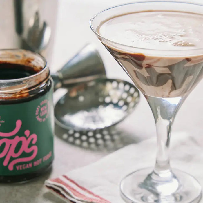 A jar of COOPS VEGAN HOT FUDGE features rich European chocolate fudge dripping down the side. The green label with pink lettering and a brown lid display the product name and description—ideal as an ice cream topping.