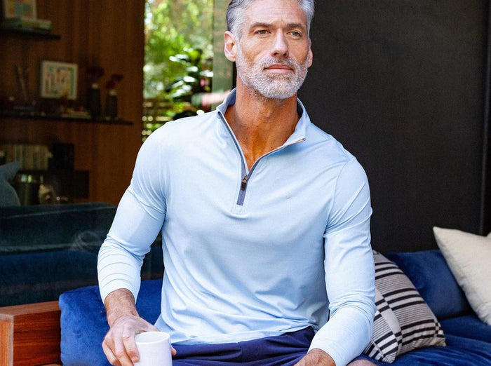 A man wears the CRIQUET Feather Performance Knit Quarter Zip Pullover in Heather Blue, featuring brrr° technology and a small embroidered logo on the chest, over a white T-shirt. His face is partially out of frame.