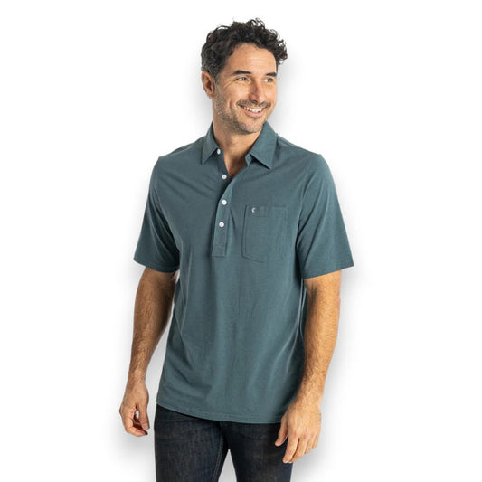 A smiling man with short dark hair wears the CRIQUET Top-Shelf Players Shirt in Dark 'n Stormy with a chest pocket and dark jeans, standing against a plain white background.