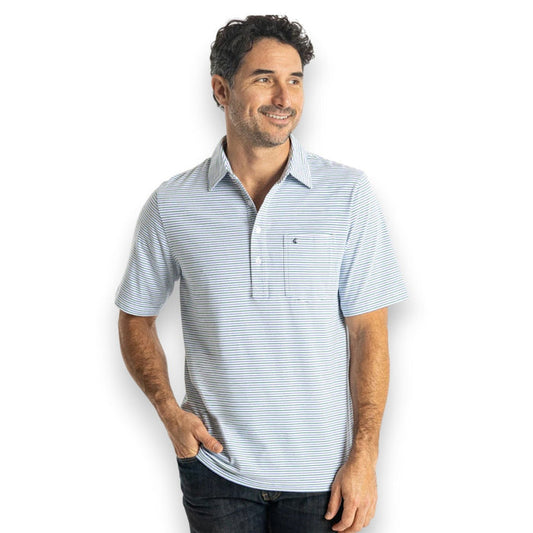 A person in sunglasses wears the CRIQUET Top-Shelf Players Shirt in Stewart Stripe White, standing by a countertop with a green can against blue-gray tiled walls.