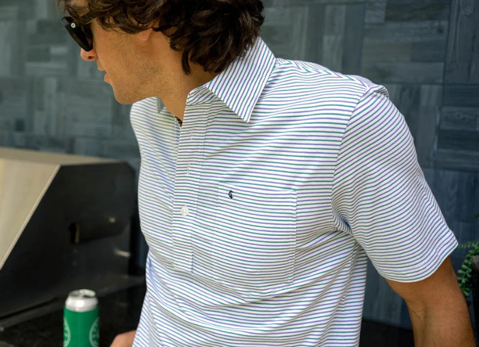 A man with dark curly hair smiles in the CRIQUET Top-Shelf Players Shirt in Stewart Stripe White, featuring light blue and white stripes, a chest pocket, and dark jeans, standing against a white background with one hand in his pocket.