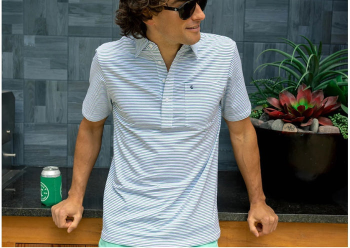A person in sunglasses wears the CRIQUET Top-Shelf Players Shirt in Stewart Stripe White, standing by a countertop with a green can against blue-gray tiled walls.