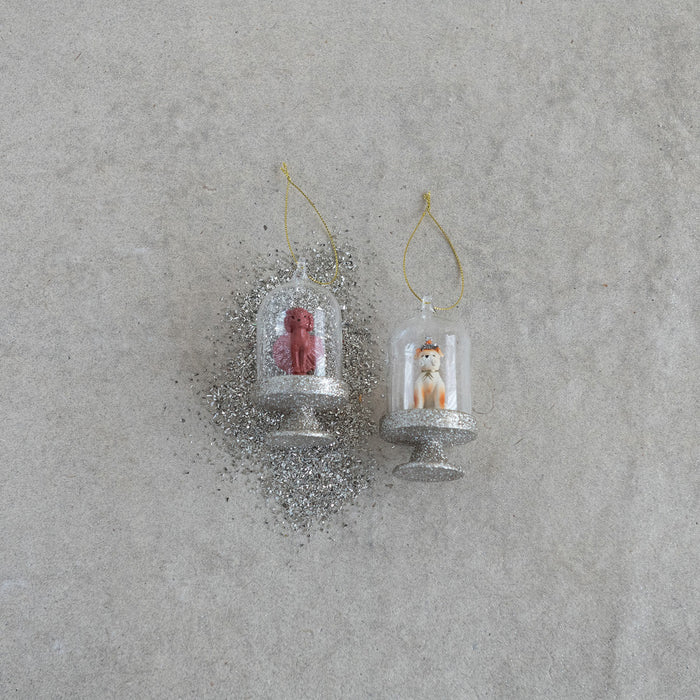 The CREATIVE COOP DOG IN CLOCHE ORNAMENT set includes two glass ornaments with gold bases and loops: one features a crowned dog, the other a pink poodle, each displayed on separate stands against a white background.