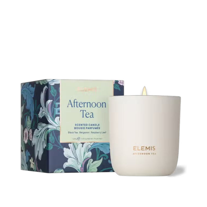 A lit ELEMIS - AFTERNOON TEA CANDLE by ELEMIS - STEINER sits in a white cup with the label "Elemis Afternoon Tea," complemented by a decorative box featuring floral designs and descriptions. The candle's scents include Black Tea, Bergamot, and Raspberry Leaf, all crafted from premium mineral wax.