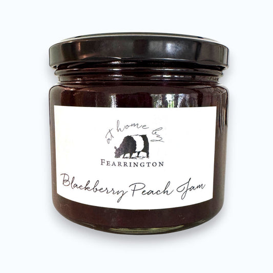 A glass jar of FEARRINGTON SIGNATURE COLLECTION - FEARRINGTON HOUSEMADE BLACKBERRY PEACH JAM with a black lid and a white label. The label lists ingredients: blackberries, peaches, lemon, cinnamon, nutmeg, sugar, pectin. It also mentions: "Handmade with care in our kitchens by Fearrington chefs." Perfect as a hostess gift or for breakfast jams. May contain allergens.
