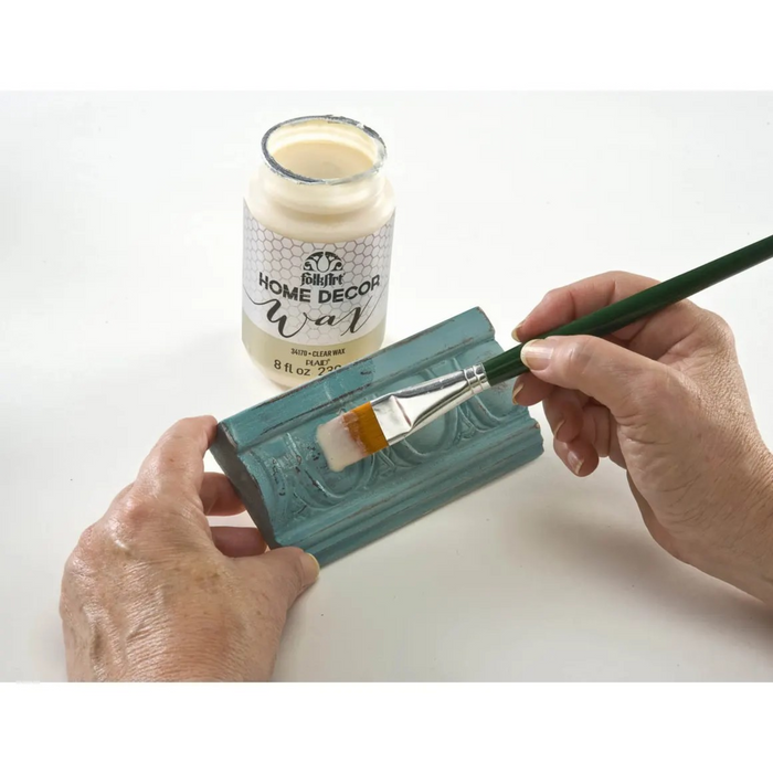 Hands applying a teal decorative wax finish to a rectangular block with a paintbrush, with a jar of Scentsy Home Decor Wax in the background on a white surface.
