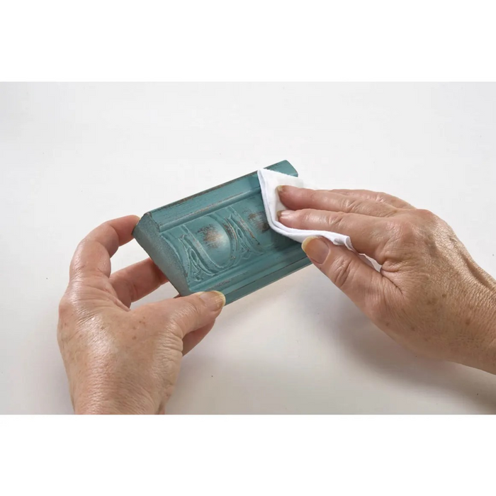 A pair of hands holding a teal rectangular decorative block or tile while wiping it with a white cloth on a plain white background.