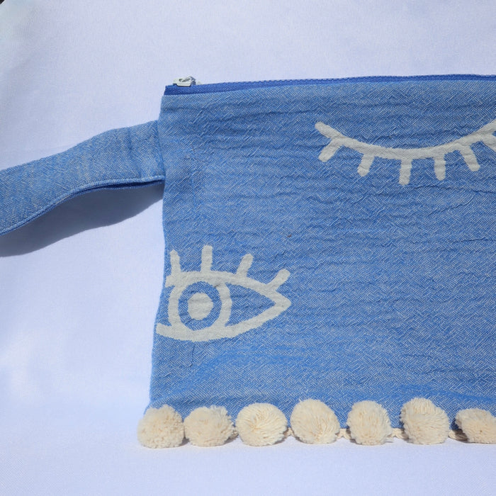 Blue clutch bag zippered wet/dry pouch with evil eye print and cream pom pom trim, made in Greece. Greek-inspired beach accessories