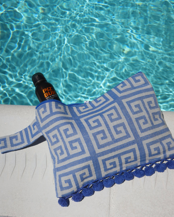 Blue and white Greek key print wet/dry clutch bag with a blue pom pom trim next to a swimming pool. Water-resistant beach bag