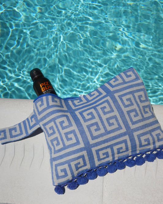 Blue and white Greek key print wet/dry clutch bag with a blue pom pom trim next to a swimming pool. Water-resistant beach bag