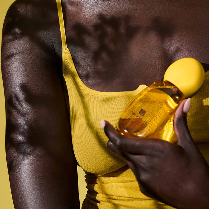 A close-up lifestyle shot of a model with dark skin wearing a yellow ribbed tank top and holding a yellow bottle of Her Wisdom Sim Sim Perfume Mist.
