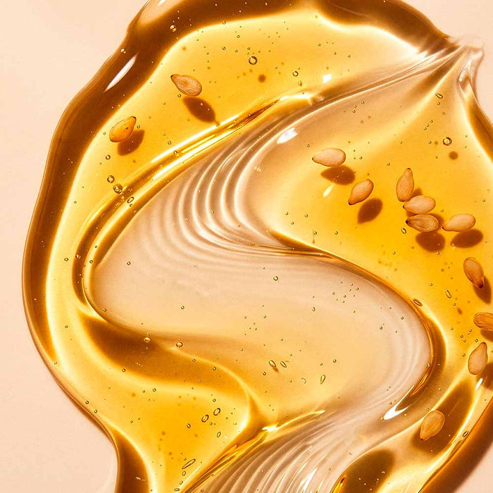 A close-up of Sim Sim Bath & Shower Oil, showing a glossy, translucent golden liquid with scattered small brown seeds on a warm beige background.
