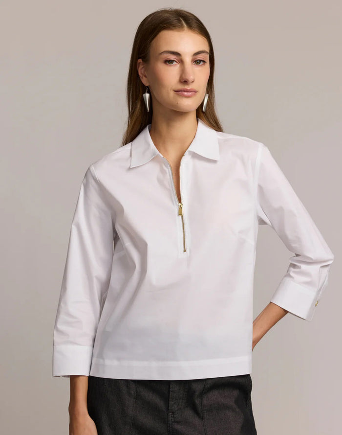 A woman stands against a plain, light background wearing the HINSON WU Alessa 3/4 Sleeve Shirt in stretch polished cotton—a crisp white blouse with a collared neckline and partial front zipper—paired with dark pants.
