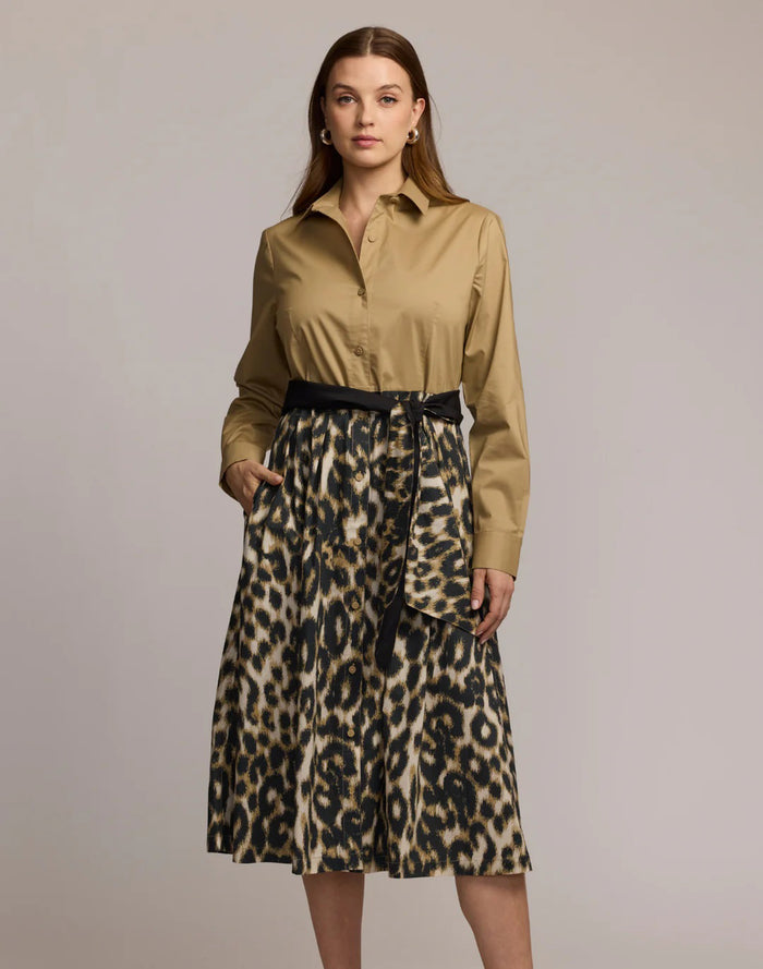 A woman stands facing forward wearing a tan long-sleeve button-up shirt tucked into a knee-length leopard print skirt with a black belt, against a plain light gray background.