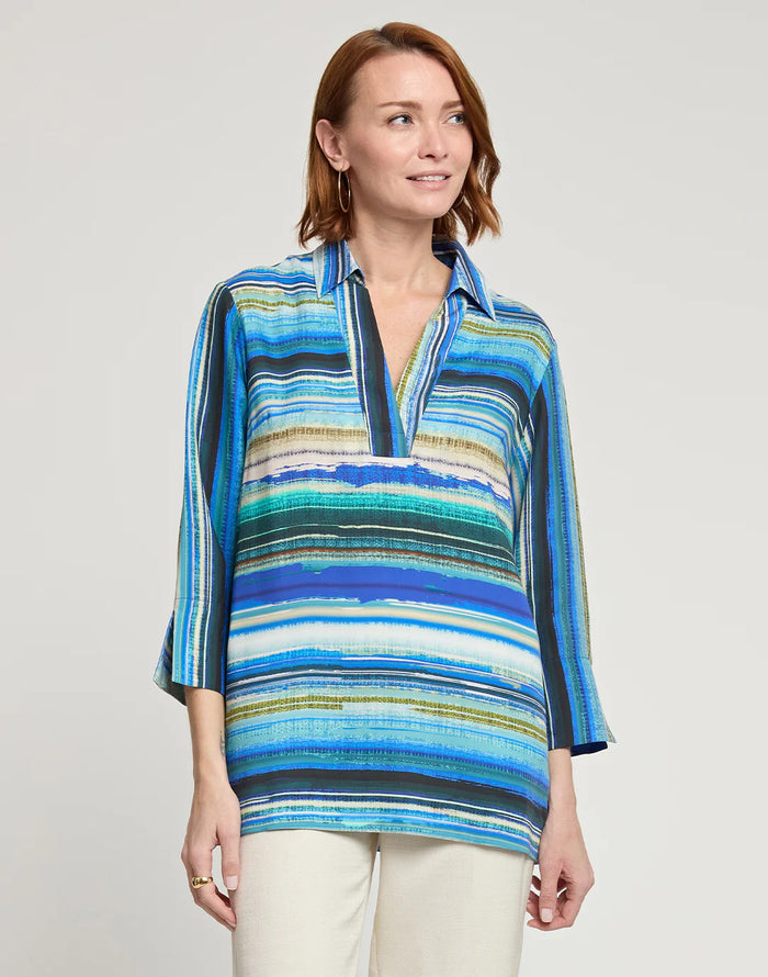 A woman wearing the HINSON WU VICKY 3/4 Sleeve Tencel Textured Stripe Print Top in blue, green, and white stripes with off-white pants stands smiling against a plain background, her light brown hair styled with hoop earrings.