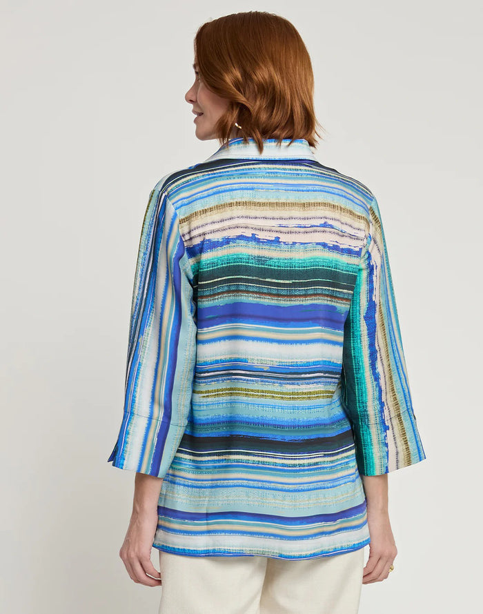A woman wearing the HINSON WU VICKY 3/4 Sleeve Tencel Textured Stripe Print Top in blue, green, and white stripes with off-white pants stands smiling against a plain background, her light brown hair styled with hoop earrings.