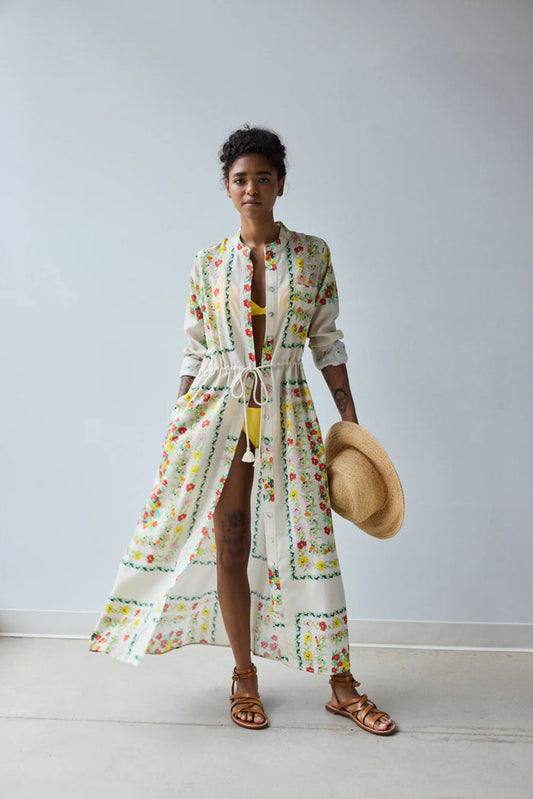 A person stands facing away in the HOPE FOR FLOWERS Band Collar Shirtdress in Custard Floral, paired with brown sandals and holding a straw hat, against a plain light gray background.