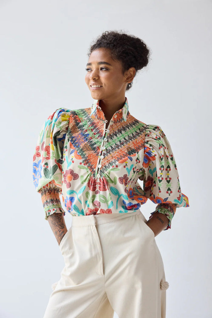 A person with curly hair wears the HOPE FOR FLOWERS Smocked Shirt in Custard Floral and cream pants, holding a straw hat at their side while glancing over their shoulder against a plain, light background.