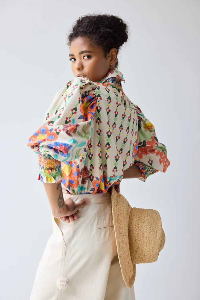 A woman smiles and looks to the side, hands in her cream pants pockets, wearing the HOPE FOR FLOWERS Smocked Shirt in Custard Floral by HOPE FOR FLOWERS against a plain light background.