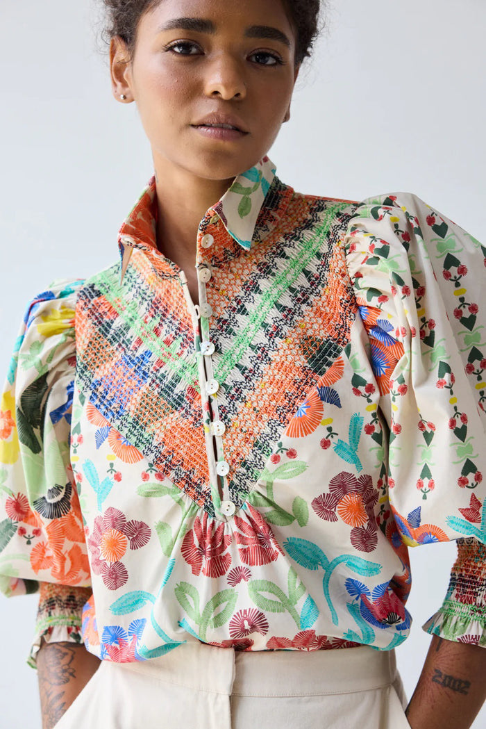 A person with curly hair wears the HOPE FOR FLOWERS Smocked Shirt in Custard Floral and cream pants, holding a straw hat at their side while glancing over their shoulder against a plain, light background.