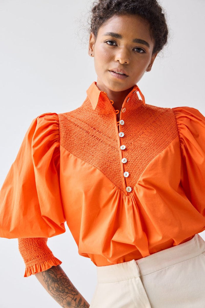 A person with curly hair is seen from behind wearing the HOPE FOR FLOWERS - SMOCKED SHIRT IN FLAME by HOPE FOR FLOWERS—an orange organic cotton poplin shirt with puff sleeves—and light pants. A numbered tattoo is visible on their neck against a plain background.