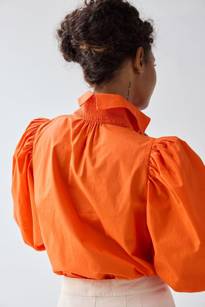 A person with curly hair is seen from behind wearing the HOPE FOR FLOWERS - SMOCKED SHIRT IN FLAME by HOPE FOR FLOWERS—an orange organic cotton poplin shirt with puff sleeves—and light pants. A numbered tattoo is visible on their neck against a plain background.