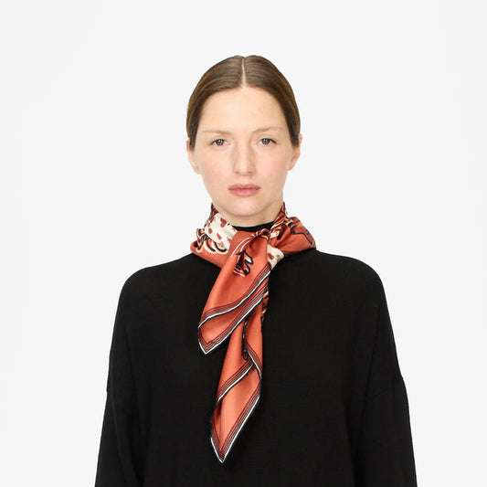 A woman with straight brown hair, tied back, wearing a black top and a shiny silk scarf in shades of orange and white, tied in a loose knot around her neck, stands against a plain white background.