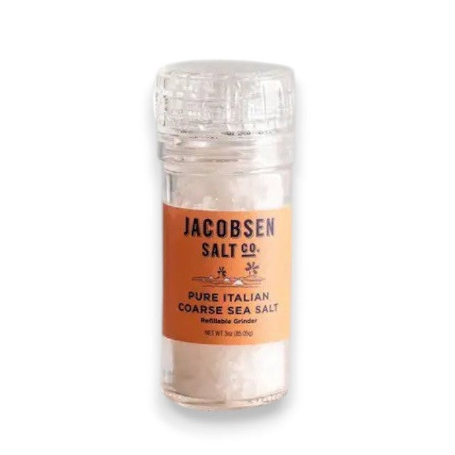 A clear plastic grinder of JACOBSEN SALT CO. JACOBSEN SALT - COARSE ITALIAN SEA SALT WITH GRINDER, sourced from the renowned Trapani region, rests on a light stone surface against a plain gray background.