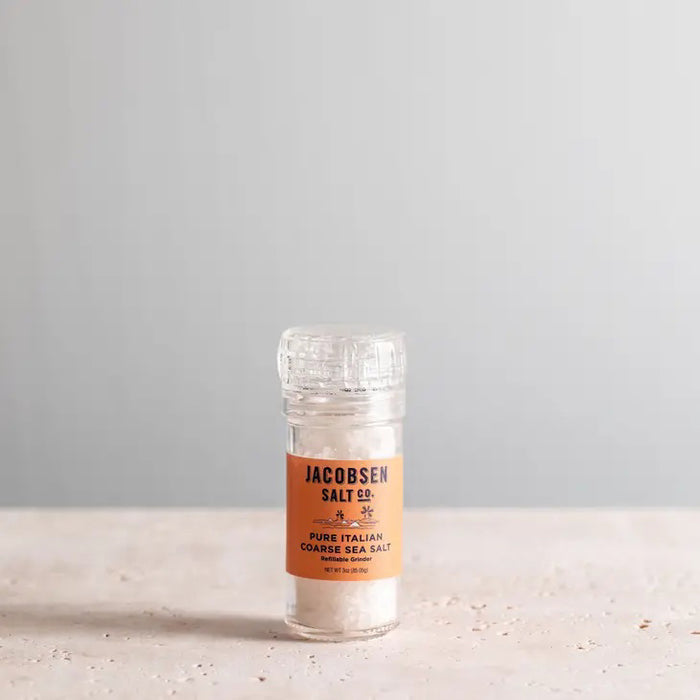 A clear plastic grinder of JACOBSEN SALT CO. JACOBSEN SALT - COARSE ITALIAN SEA SALT WITH GRINDER, sourced from the renowned Trapani region, rests on a light stone surface against a plain gray background.