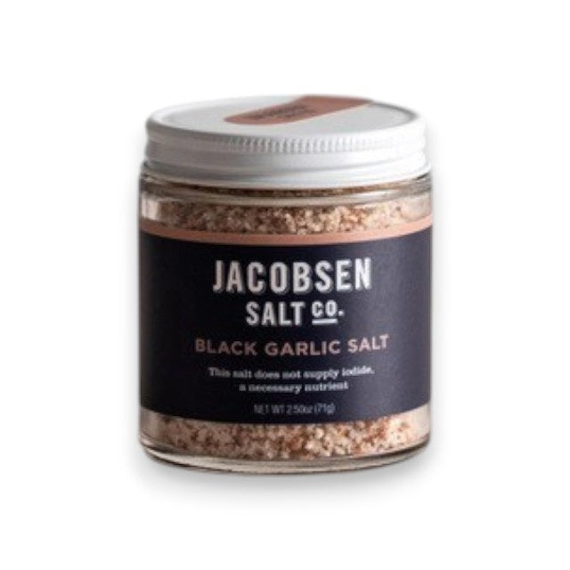 A jar of JACOBSEN SALT CO. INFUSED BLACK GARLIC SALT, featuring a black label and white lid, rests on a light beige surface against a plain light gray background.