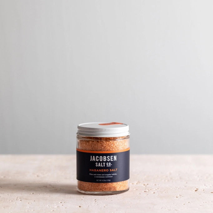 A small jar of JACOBSEN SALT CO. JACOBSEN SALT - INFUSED HABANERO SALT with a white lid sits on a light-colored surface against a plain, light gray background, ideal as spicy salt or for rimming cocktails.