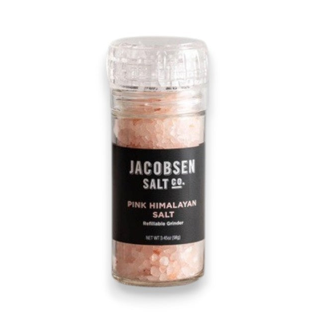 A transparent glass grinder containing JACOBSEN SALT - PINK HIMALAYAN SALT rests on a light surface. Featuring a black label with the brand name “JACOBSEN SALT CO.” and the product description, this refillable grinder is positioned against a simple, light gray background.