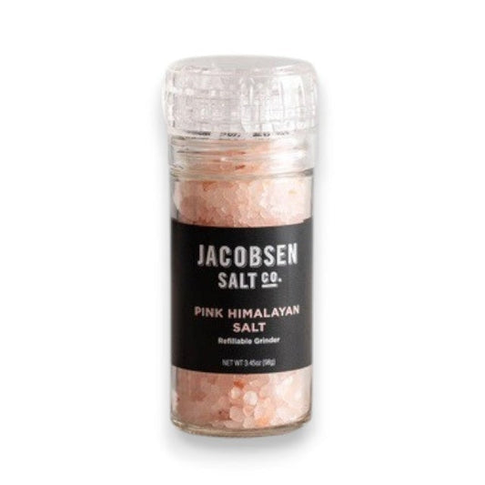 A transparent glass grinder containing JACOBSEN SALT - PINK HIMALAYAN SALT rests on a light surface. Featuring a black label with the brand name “JACOBSEN SALT CO.” and the product description, this refillable grinder is positioned against a simple, light gray background.