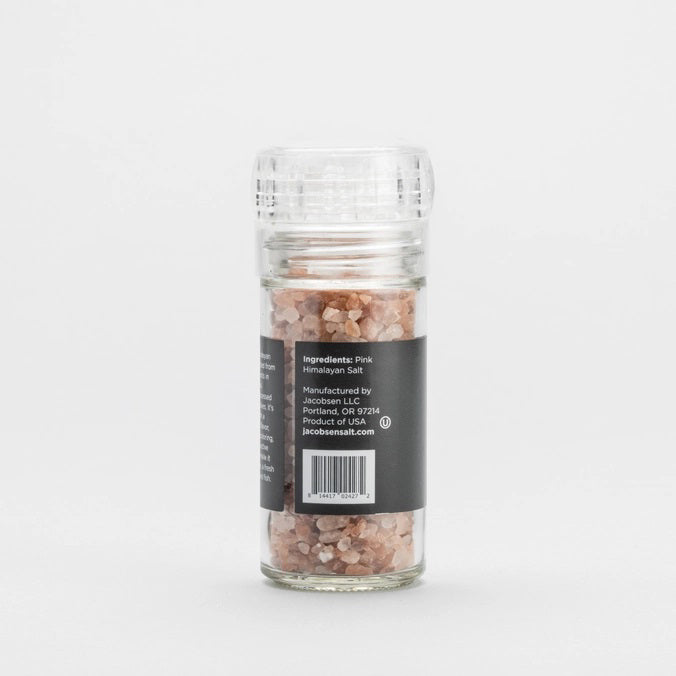 A transparent glass grinder containing JACOBSEN SALT - PINK HIMALAYAN SALT rests on a light surface. Featuring a black label with the brand name “JACOBSEN SALT CO.” and the product description, this refillable grinder is positioned against a simple, light gray background.