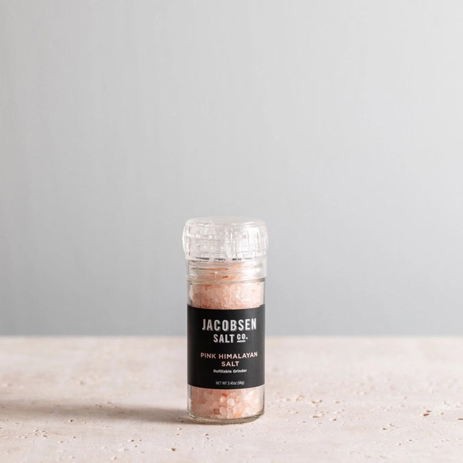 A transparent glass grinder containing JACOBSEN SALT - PINK HIMALAYAN SALT rests on a light surface. Featuring a black label with the brand name “JACOBSEN SALT CO.” and the product description, this refillable grinder is positioned against a simple, light gray background.