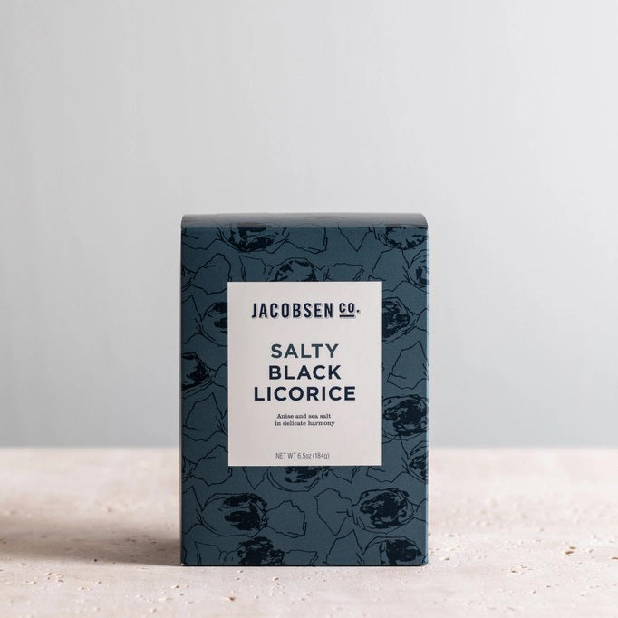 A dark blue JACOBSEN SALT - SALTY BLACK LICORICE BOX by JACOBSEN SALT CO. rests on a light surface against a plain gray background. Its white label with black text and abstract patterns hints at the sea salt and anise flavors inside.