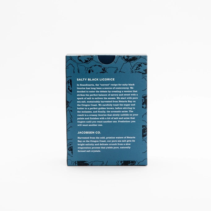A dark blue JACOBSEN SALT - SALTY BLACK LICORICE BOX by JACOBSEN SALT CO. rests on a light surface against a plain gray background. Its white label with black text and abstract patterns hints at the sea salt and anise flavors inside.