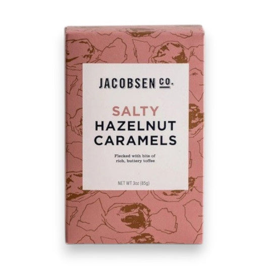 A pink JACOBSEN SALT - SALTY HAZELNUT CARAMELS BOX by JACOBSEN SALT CO. sits on a light textured surface, featuring a modern design with subtle hazelnut patterns and hints of buttery toffee made from premium hazelnuts.