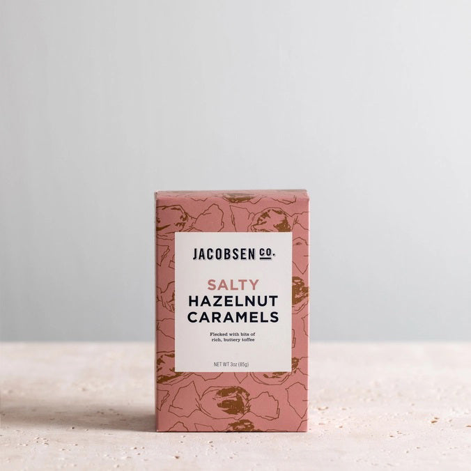 A pink JACOBSEN SALT - SALTY HAZELNUT CARAMELS BOX by JACOBSEN SALT CO. sits on a light textured surface, featuring a modern design with subtle hazelnut patterns and hints of buttery toffee made from premium hazelnuts.