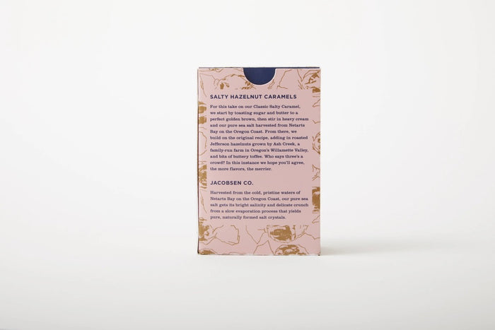 A pink JACOBSEN SALT - SALTY HAZELNUT CARAMELS BOX by JACOBSEN SALT CO. sits on a light textured surface, featuring a modern design with subtle hazelnut patterns and hints of buttery toffee made from premium hazelnuts.