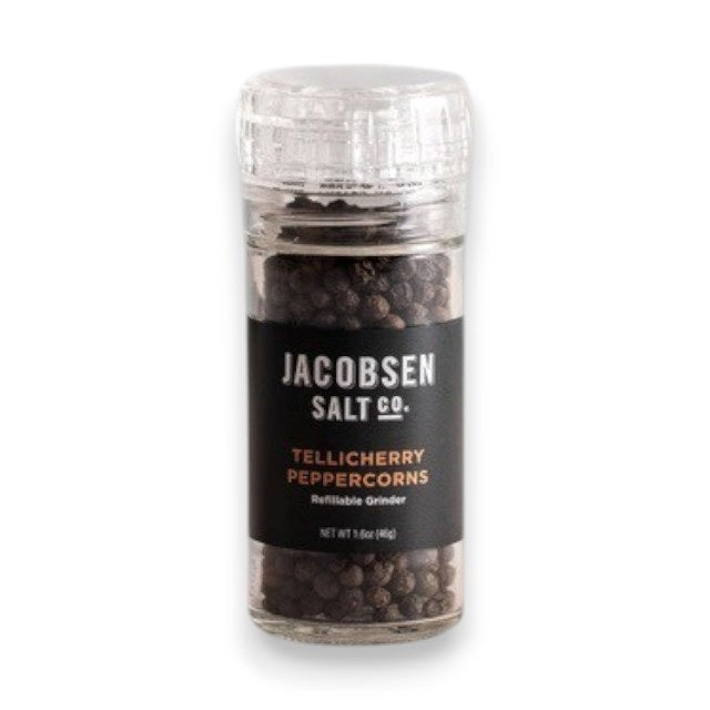 A clear glass grinder filled with black peppercorns rests on a light surface. The label displays “Jacobsen Salt Co.” and “Tellicherry Peppercorns” in white and gold text. Behind, a plain light gray wall forms the background. This is the JACOBSEN SALT - TELLICHERRY PEPPERCORNS WITH GRINDER by JACOBSEN SALT CO.
