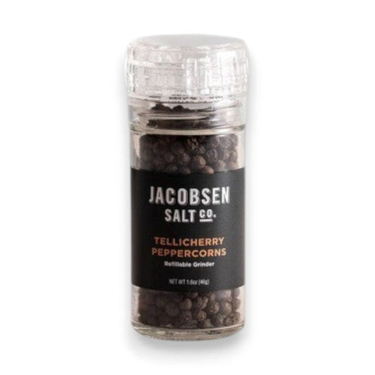 A clear glass grinder filled with black peppercorns rests on a light surface. The label displays “Jacobsen Salt Co.” and “Tellicherry Peppercorns” in white and gold text. Behind, a plain light gray wall forms the background. This is the JACOBSEN SALT - TELLICHERRY PEPPERCORNS WITH GRINDER by JACOBSEN SALT CO.