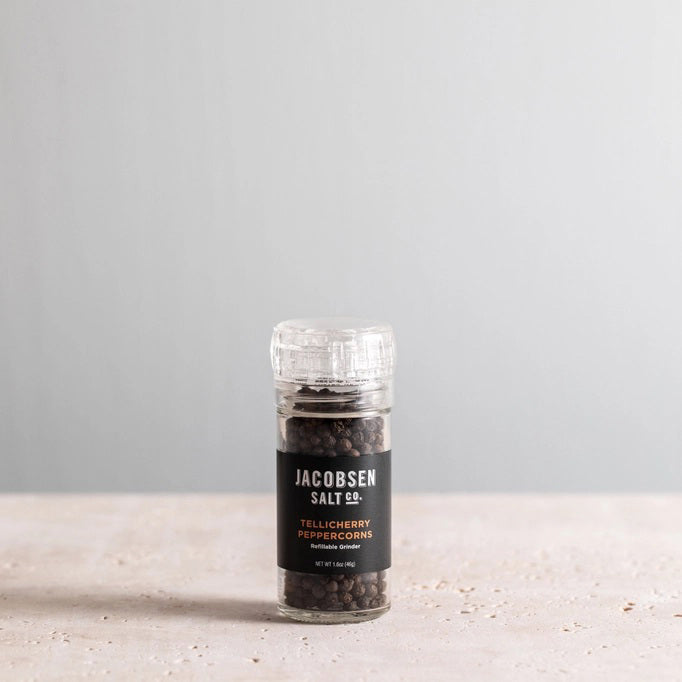 A clear glass grinder filled with black peppercorns rests on a light surface. The label displays “Jacobsen Salt Co.” and “Tellicherry Peppercorns” in white and gold text. Behind, a plain light gray wall forms the background. This is the JACOBSEN SALT - TELLICHERRY PEPPERCORNS WITH GRINDER by JACOBSEN SALT CO.