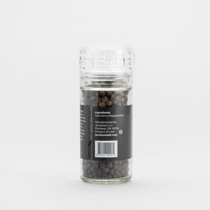 A clear glass grinder filled with black peppercorns rests on a light surface. The label displays “Jacobsen Salt Co.” and “Tellicherry Peppercorns” in white and gold text. Behind, a plain light gray wall forms the background. This is the JACOBSEN SALT - TELLICHERRY PEPPERCORNS WITH GRINDER by JACOBSEN SALT CO.