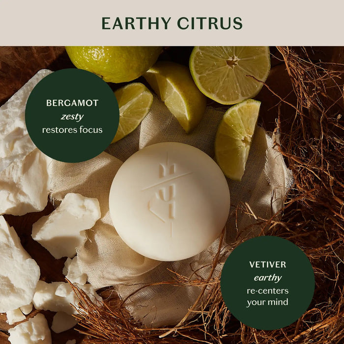 A round white KATE MCLEOD Grounding Stone deep moisturizing lotion bar sits on textured fabric next to an open container with a wooden lid, featuring a dark green label with orange and white text, highlighting its luxurious blend of cocoa butter concentrate and natural essential oils.
